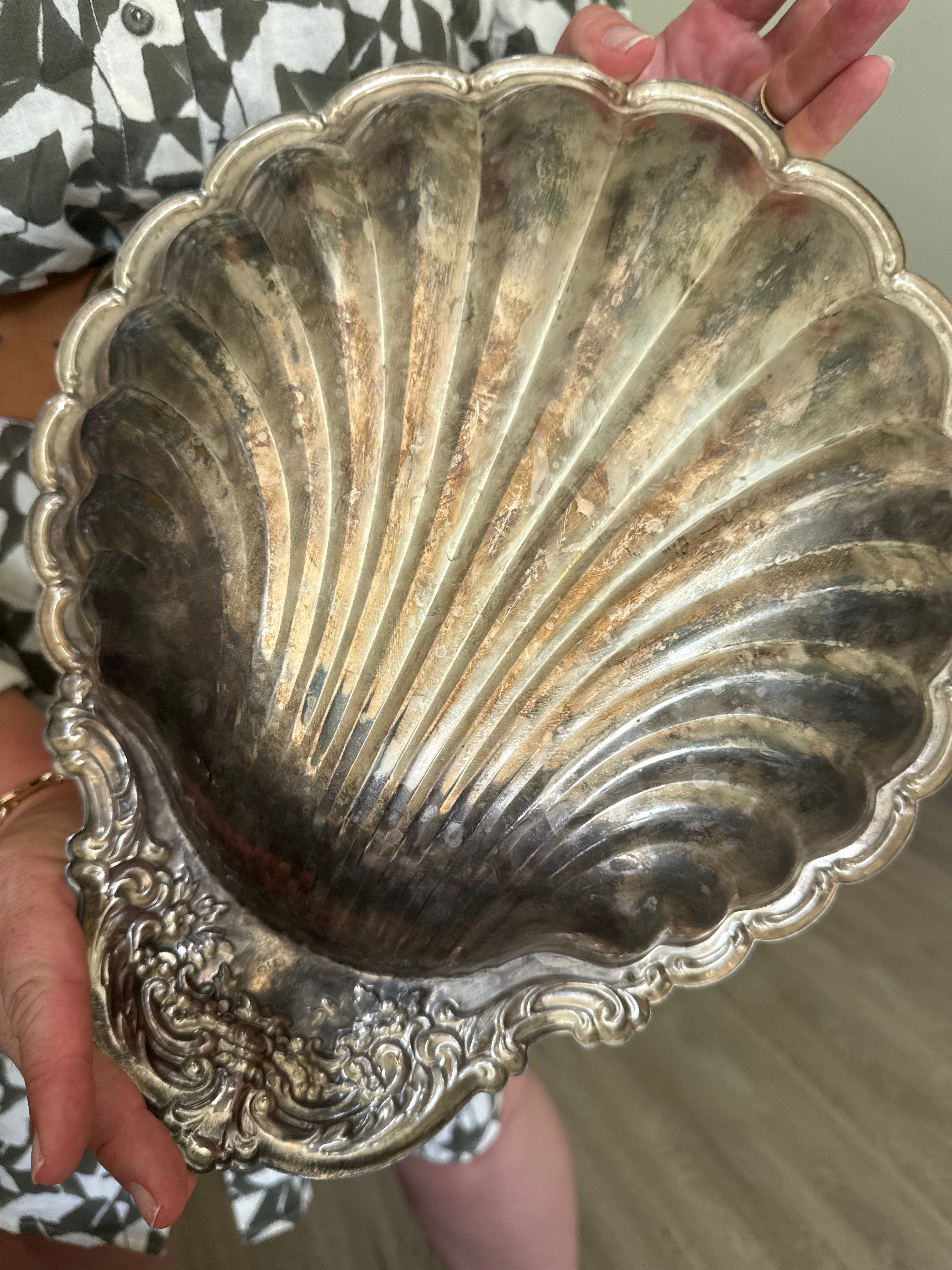 A vintage silver shell-shaped serving platter with an ornate carved base, held by a person in a black and white patterned shirt.