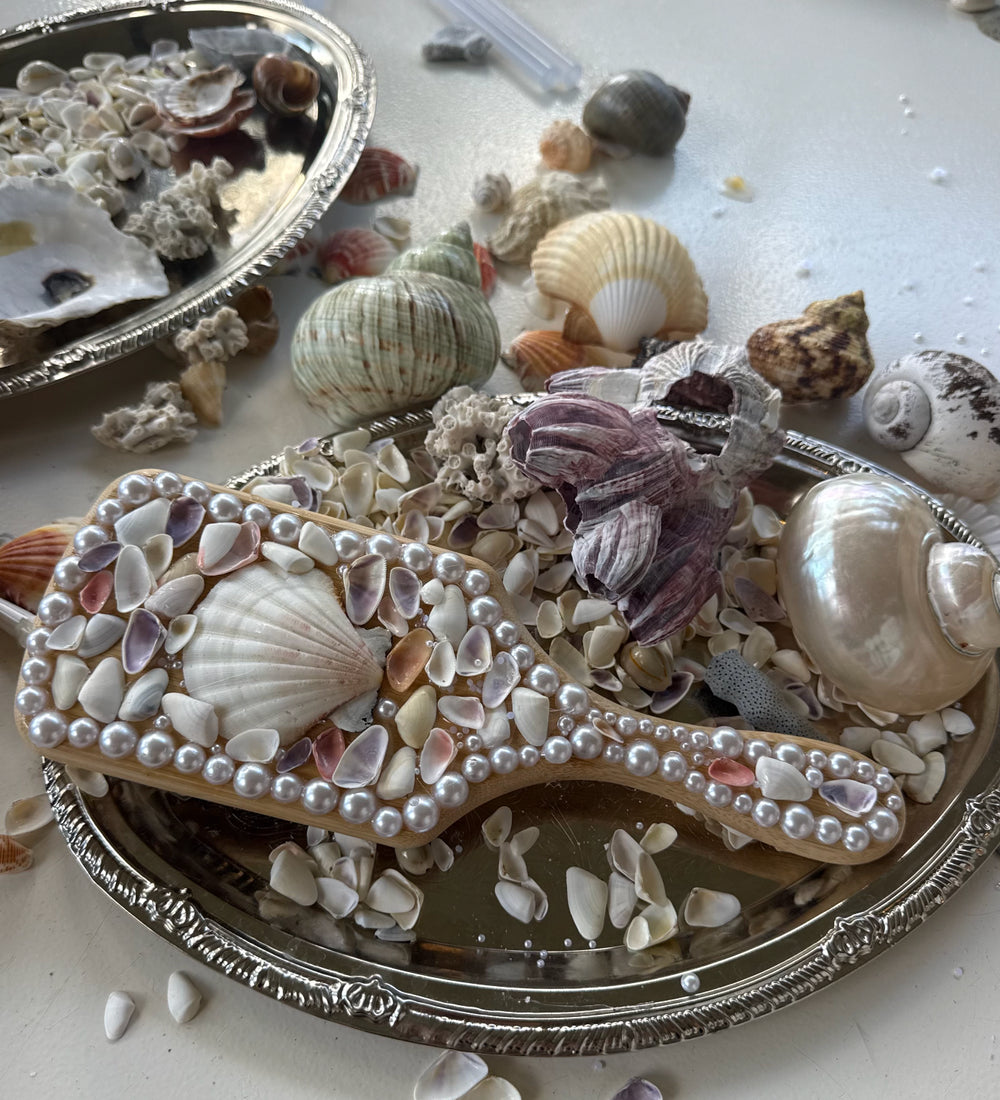 A decorative wooden hair comb embellished with white faux pearls and seashells, displayed on an ornate silver tray surrounded by various real seashells.