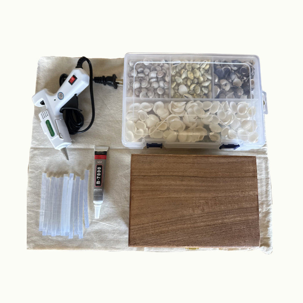 A DIY craft kit laid out on a light beige fabric background, including a hot glue gun with its cord, a clear plastic organizer filled with small decorative pieces, a tube of adhesive, a bundle of clear plastic fasteners, and a rectangular wooden board.