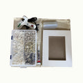 A DIY craft kit laid out on a light beige fabric surface, including a white hot glue gun, a clear plastic organizer filled with decorative shells, a small tube of adhesive, a clear plastic bag, and a white wooden frame with a square cutout.