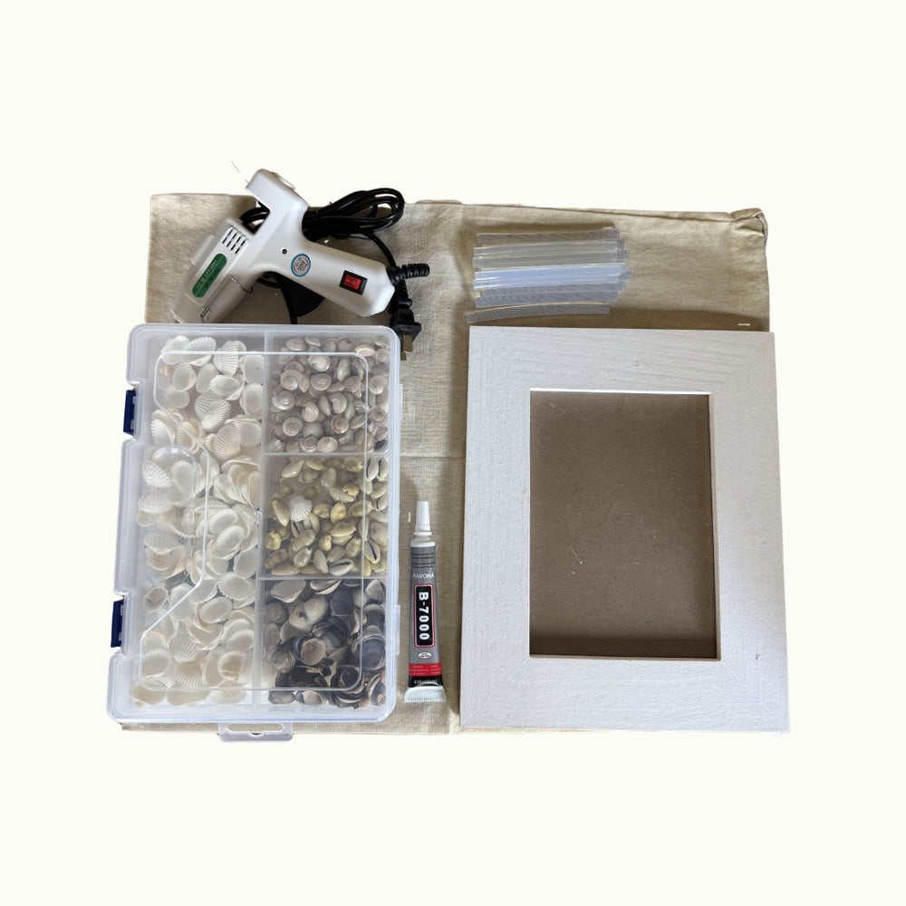 A DIY craft kit laid out on a light beige fabric surface, including a white hot glue gun, a clear plastic organizer filled with decorative shells, a small tube of adhesive, a clear plastic bag, and a white wooden frame with a square cutout.